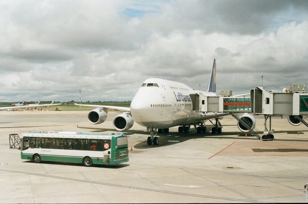 pexels-photo-11524258 A large airplane docked at an airport gate with a shuttle bus nearby under cloudy skies.